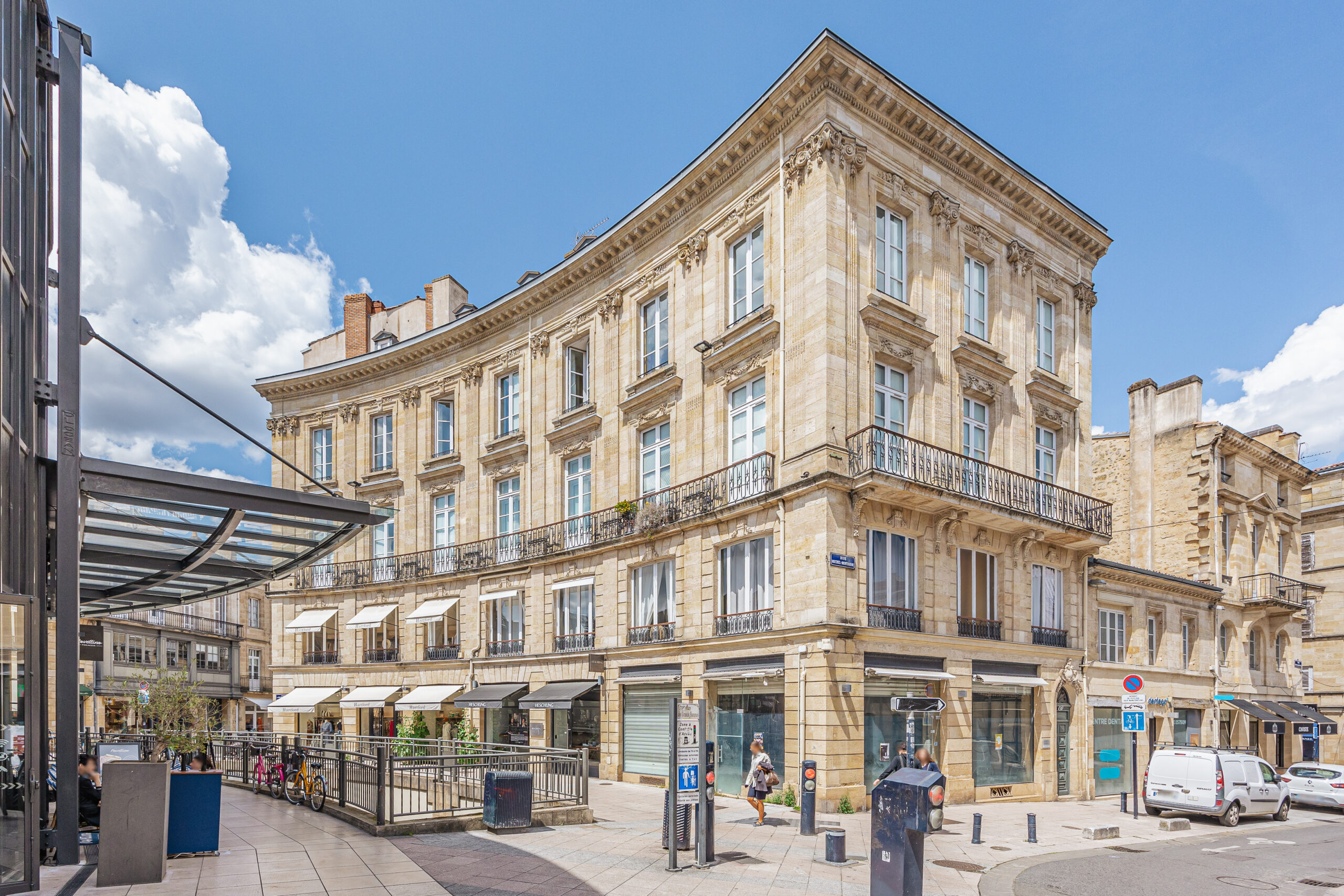 Superbe immeuble en pierre de Bordeaux situé en centre-ville, avec façades élégantes, balcons en fer forgé et boutiques en rez-de-chaussée, sous un ciel bleu ensoleillé.